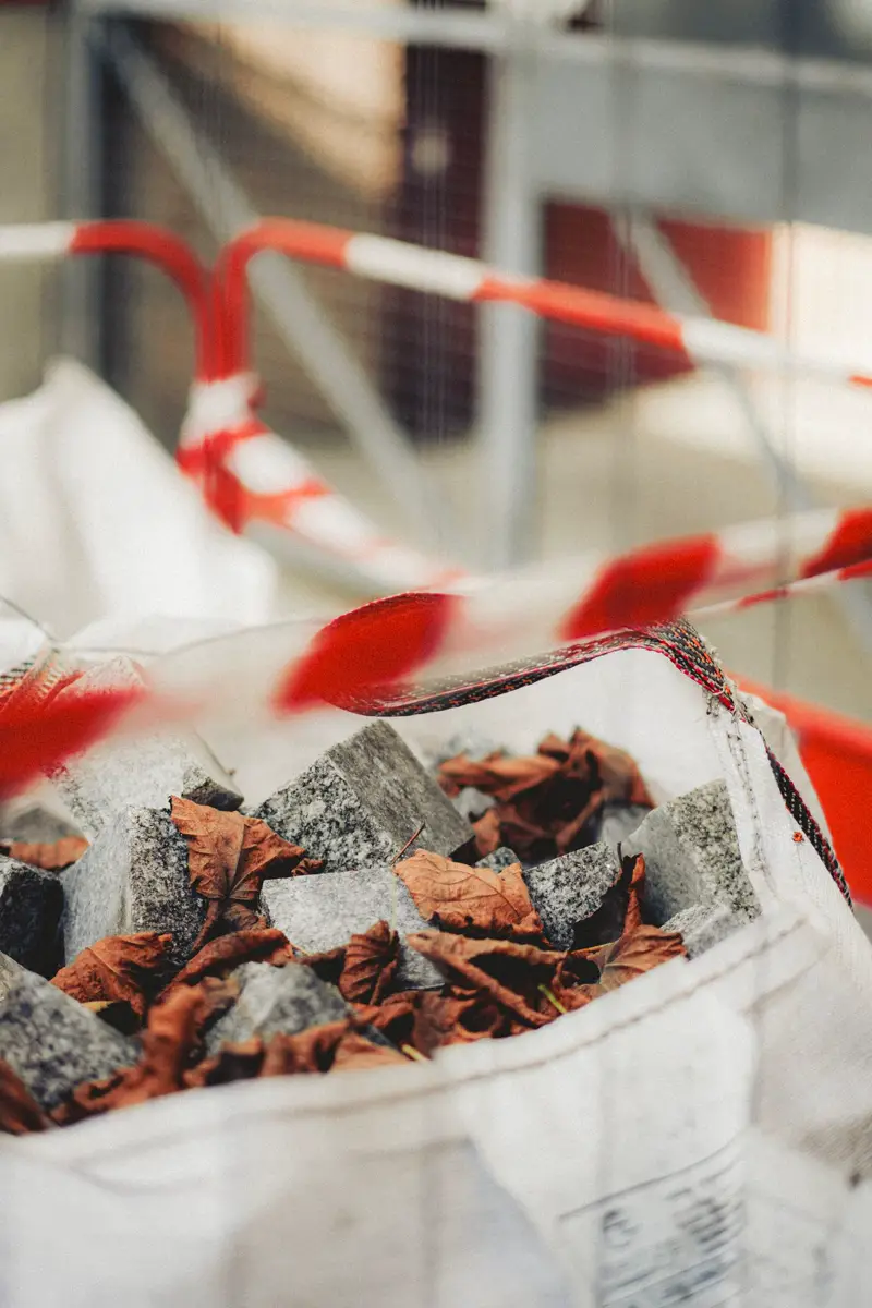A white woven dumpster bag filled with grey stone blocks and dry leaves at a work site.