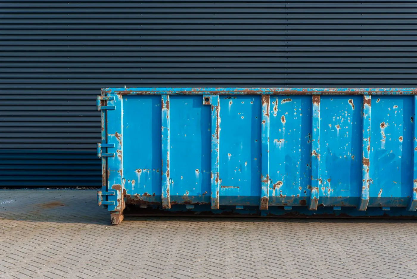 An open-top steel roll-off dumpster on brick pavers, with rusted ribs and a rear swinging door. No yardage is marked.